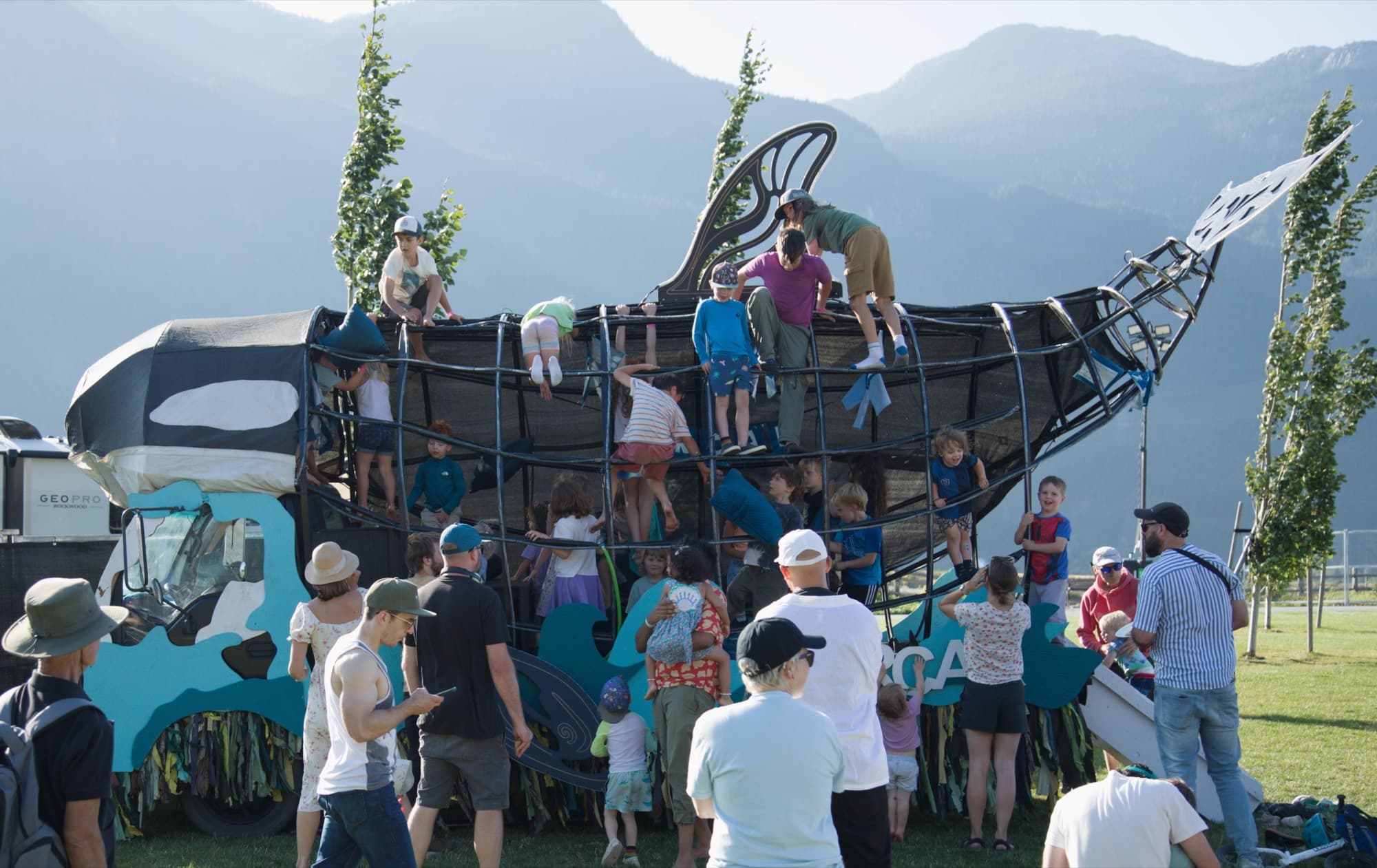 The same Orcar in afternoon sun, kids climbing all over its steel ribs and dorsal fin, the Squamish mountains hazy behind