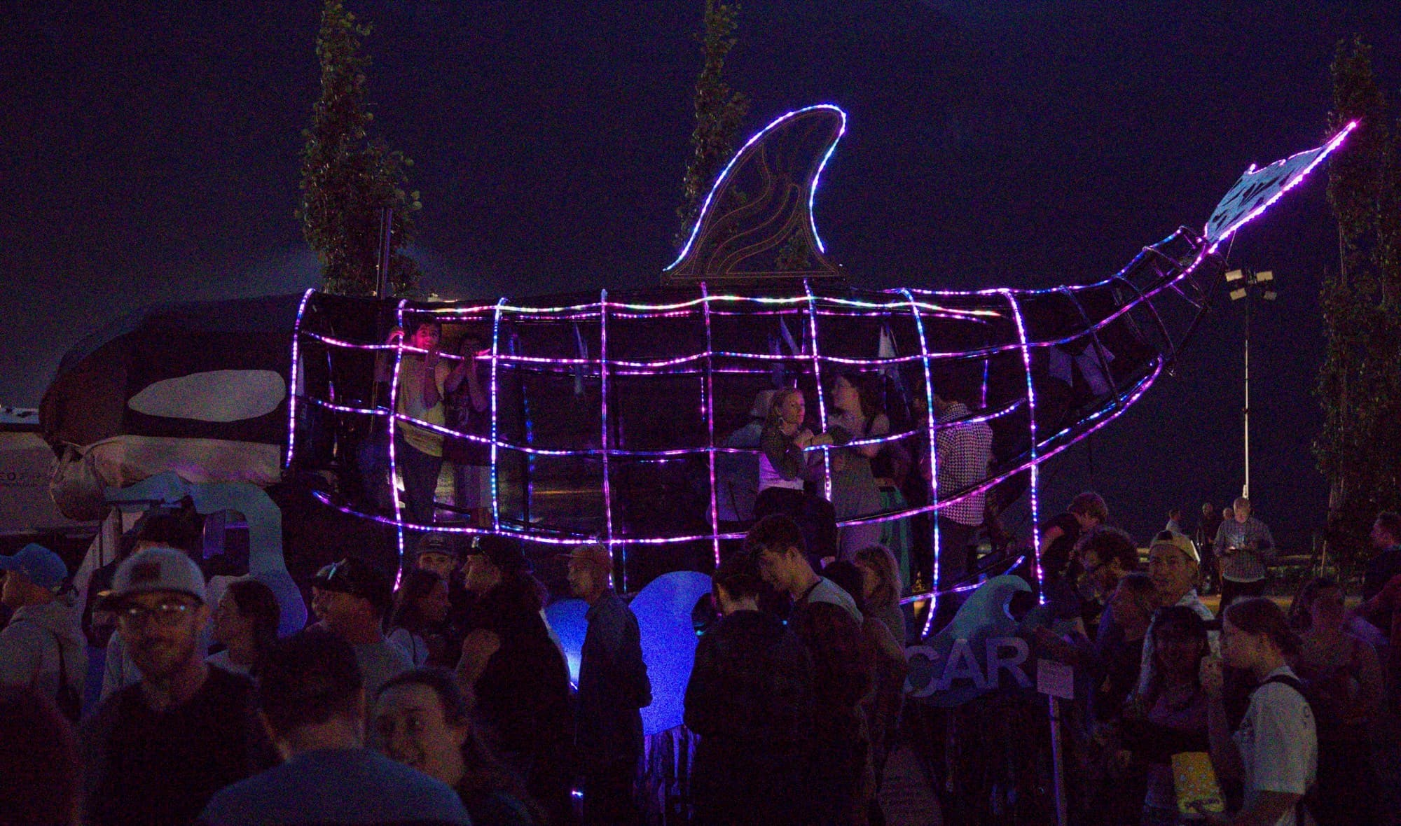 The Orcar at night lit in pink and blue LED rope, riders inside the ribs above a dense festival crowd