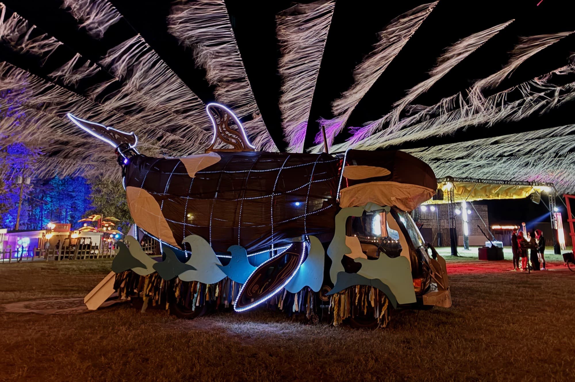 The Orcar parked at night under a huge fringed canopy, its body and waves outlined in white LED, a lit stage and market stalls glowing behind
