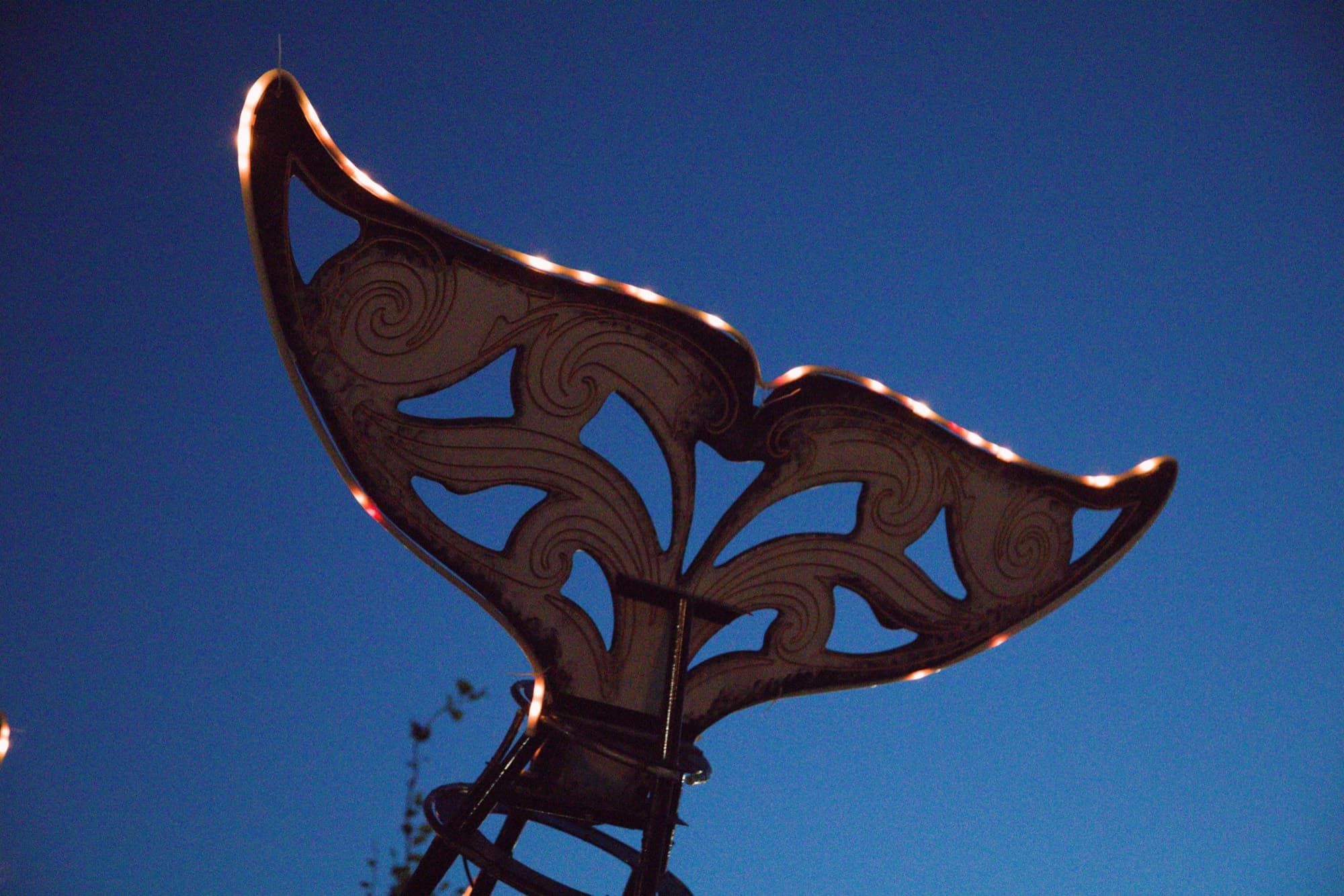 The Orcar's laser-cut tail fluke against a deep blue twilight sky, its carved wave curls edged in warm LED light